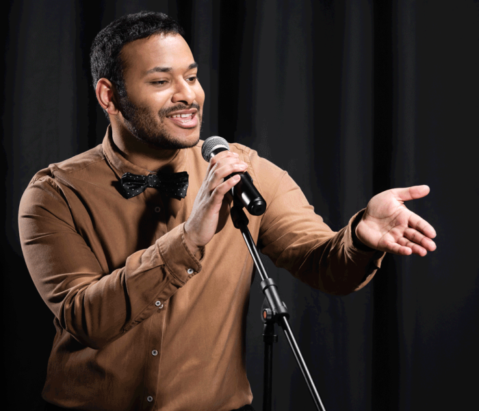 indian comedian performing stand up comedy and telling jokes in microphone while gesturing on black, banner. 