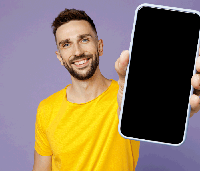 Young fun man wear yellow t-shirt hold in hand use show close up mobile cell phone with blank screen workspace area isolated on plain pastel light purple